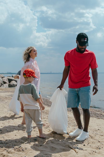 A group of three individuals participating in a beach clean-up activity during daytime, with an open sky filled with mostly cloudy weather. The scene features a young woman with curly blonde hair standing to the left, dressed in a light pink top, looking towards the water in the background. To her right, a small boy wearing a red cap with a pattern, a grey and white shirt, and light grey shorts is holding a white rubbish bag. Adjacent to the boy, a man dressed in a red T-shirt, light blue denim shorts, white sneakers, and a black cap with a small logo is also holding a white rubbish bag, actively engaged in collecting waste from the sandy shoreline. Behind them, there is a calm body of water with a long stretch of rocky shoreline, and the overall environment appears to be a natural coastal area, where private waste disposal methods or independent rubbish collection could be involved. The scene emphasizes community participation in rubbish removal, supported by local waste management services such as Rubbish Removal Brixton, to promote cleaner outdoor spaces.