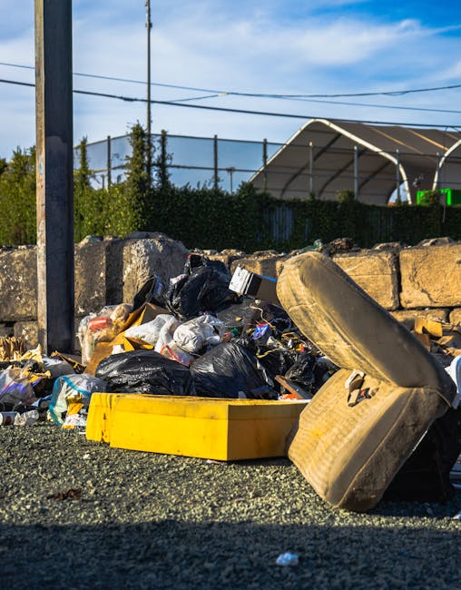 A low-angle view of a pile of mixed rubbish including black plastic garbage bags, cardboard boxes, and discarded household items, scattered across a rough asphalt surface next to a short stone wall and metal pole. In the foreground, a worn, beige car seat with visible stains and frayed fabric is partially resting on the debris. Behind the debris, a row of dense green hedges runs parallel to a fence, with an outdoor sports court or covered area visible in the background under a partly cloudy blue sky. The scene suggests an area prepared for independent waste collection or private rubbish removal, often managed by services like Rubbish Removal Brixton, rather than municipal disposal.