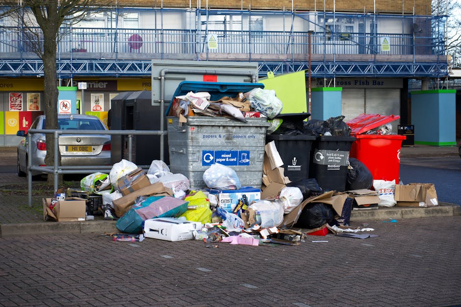 A large collection of mixed waste and rubbish is piled up beside a grey recycling bin labeled for paper and cardboard, situated on a paved urban street outside a commercial building. The rubbish includes various cardboard boxes, plastic bags, paper packaging, and loose papers spilling onto the ground. The garbage appears haphazardly stacked, with some items leaning against the bin and others scattered in front of it. Nearby, a silver car is parked parallel to the sidewalk, partially obstructed by a metal railing. Behind the pile, the building features a blue scaffolding structure covering the upper level, with storefronts on the ground floor, including signs for a fish bar and other shops. The scene is illuminated by daylight, with a large tree to the left providing some partial shade. This image illustrates an instance of on-site rubbish accumulation on a public area, highlighting the importance of private waste removal services like those provided by Rubbish Removal Brixton for maintaining tidy urban environments.