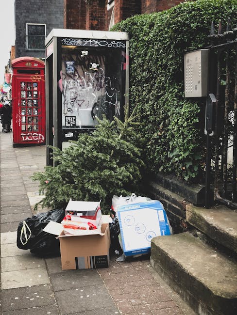 On a paved sidewalk area near a black metal fence and richly foliaged green hedge, there is a collection of discarded household rubbish and a small, green pine tree. The waste includes a cardboard box with some red and white packaging material inside, a blue and white cardboard box with a circular pattern, and a black plastic bag. The rubbish is placed directly on the ground, next to a low stone step leading up to a residential entrance. Adjacent to the waste, a white rectangular rubbish bin with graffiti marks on its transparent front panel is positioned, and behind it, a bright red British postal box stands to the left. The scene is outdoors in an urban setting with a brick building visible in the background, and the lighting appears natural, suggesting daytime. This scene illustrates an example of private rubbish disposal or waste clearance, typical of independent rubbish removal services such as those offered by Rubbish Removal Brixton, operating in the Brixton SW9 area near Loughborough Estate.