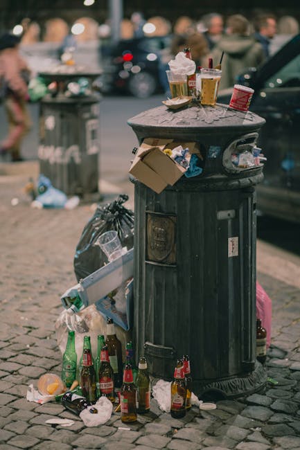 A large collection of mixed waste and rubbish is piled up beside a grey recycling bin labeled for paper and cardboard, situated on a paved urban street outside a commercial building. The rubbish includes various cardboard boxes, plastic bags, paper packaging, and loose papers spilling onto the ground. The garbage appears haphazardly stacked, with some items leaning against the bin and others scattered in front of it. Nearby, a silver car is parked parallel to the sidewalk, partially obstructed by a metal railing. Behind the pile, the building features a blue scaffolding structure covering the upper level, with storefronts on the ground floor, including signs for a fish bar and other shops. The scene is illuminated by daylight, with a large tree to the left providing some partial shade. This image illustrates an instance of on-site rubbish accumulation on a public area, highlighting the importance of private waste removal services like those provided by Rubbish Removal Brixton for maintaining tidy urban environments.