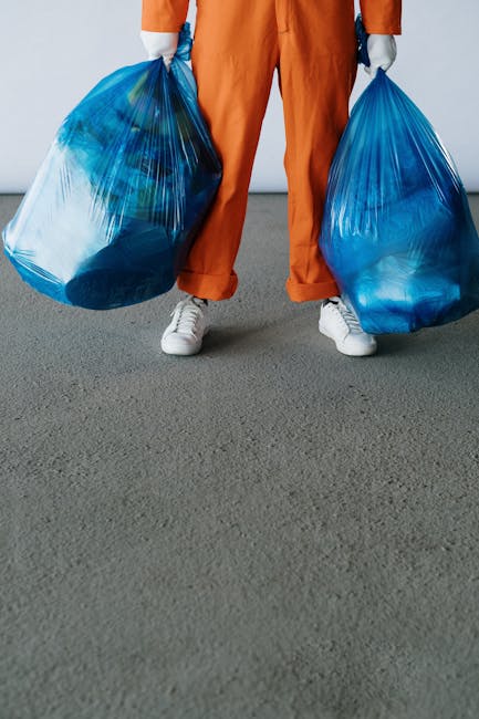 A person standing on a grey concrete surface, visible from the waist down, is holding two large blue plastic rubbish bags, one in each hand. They are wearing orange overalls and white sneakers. The rubbish bags are filled with waste materials, which can be seen through the semi-transparent plastic, and are tied at the top. The background is plain and light-colored, suggesting an indoor or outdoor setting with neutral lighting. The scene depicts an act of waste collection or removal, consistent with services offered by Rubbish Removal Brixton for efficient, private disposal and on-site clearance of rubbish. The image emphasizes manual rubbish handling and the use of plastic bags typical of private waste management processes in the context of removing waste from events or other activities requiring independent collection.