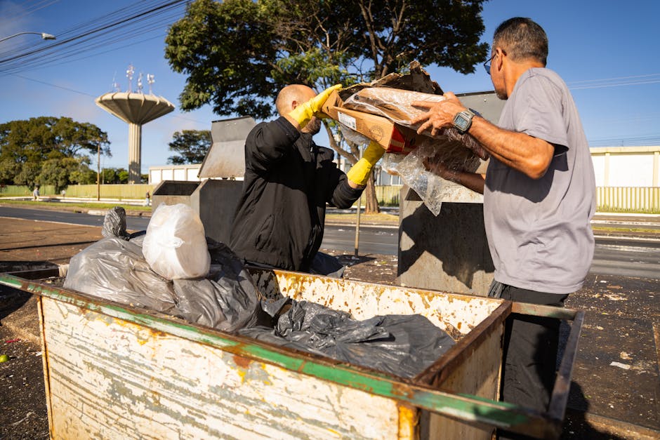 Two men engaged in rubbish collection near an open-top metal skip container on a paved outdoor area. The man on the left, wearing a black jacket and yellow gloves, is lifting a cardboard box filled with mixed waste materials, including plastic and paper, towards the skip. The man on the right, dressed in a light grey t-shirt, is holding the cardboard box steady for loading. The skip, which appears rusty and weathered, contains several black and grey plastic bags along with other debris. In the background, there are large trees with green foliage, a white building with a yellow fence, and a tall water tower visible under a clear blue sky, suggesting a spacious, open environment typical of a waste collection or disposal site. The scene is lit by natural daylight, capturing the straightforward activity of rubbish removal, aligning with services such as independent waste handling or on-site clearance provided by Rubbish Removal Brixton, without promoting any specific service.